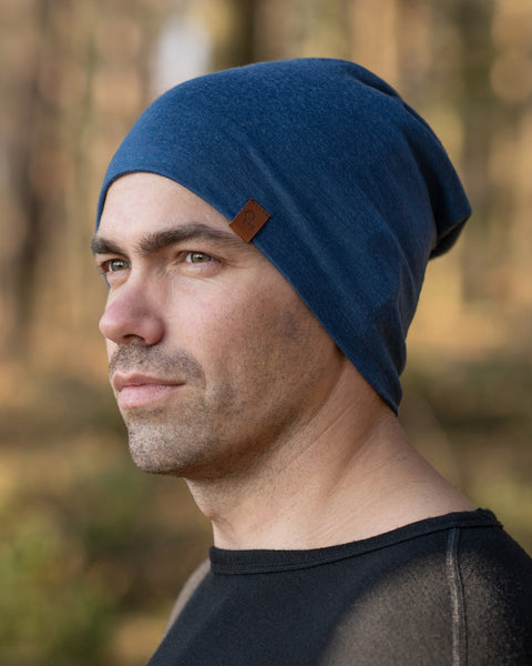 A man standing outside during fall season, in a forest, wearing the menique Men&#39;s Merino Beanie Hat in Denim Color.