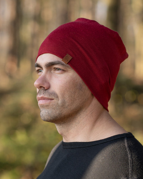 A man standing outside in a forest during fall season, wearing the menique Men&#39;s Merino Beanie Hat in Royal Cherry Color.