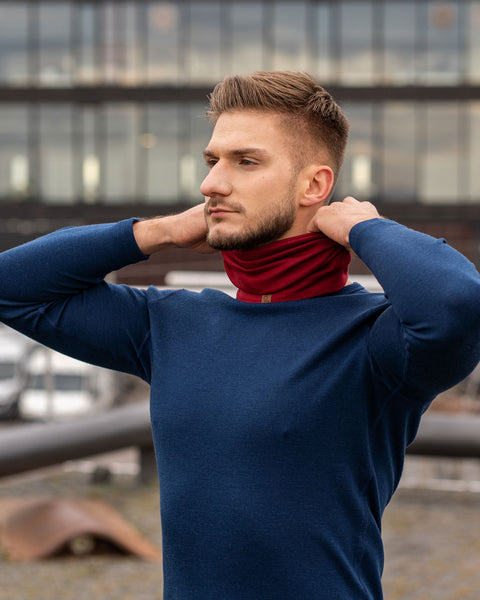 A man, outside in a city, wearing the menique unisex neck gaiter in royal cherry color. He is fixing it on his neck with his hands raised. He is also wearing a merino long sleeve top in blue color.