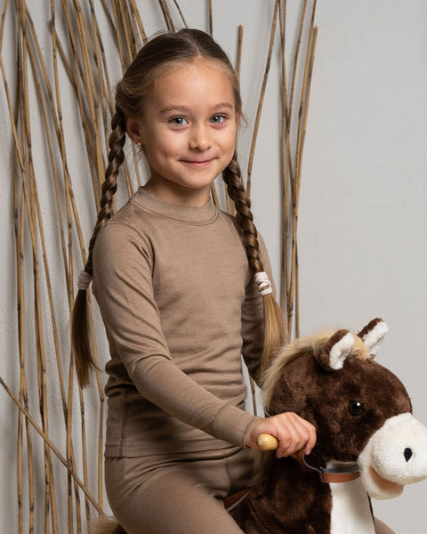A girl wearing the menique Kids&#39; Merino 160 Long Sleeve Crew in Beige color, riding a kids&#39; toy horse. 