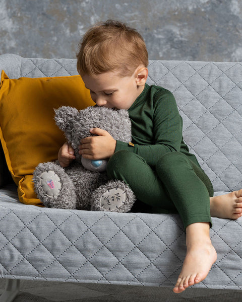 A young child with light brown hair, wearing a dark green long-sleeved shirt and matching pants, is sitting on a gray quilted sofa and hugging a gray teddy bear with a blue nose. The child&#39;s face is pressed against the bear. 