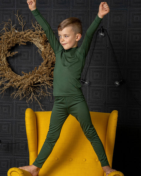 A boy wearing the menique kids&#39; 160gsm merino set in dark green color. It ncludes a long sleev etop and pants. He has his arms raised up straight and legs wide apart, standing on a spicy yellow color ottoman.
