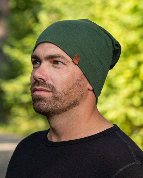 A side profile head-shot of a man wearing the Unisex Merino Beanie Hat Dark Green. Behind him is a blurred view of trees.